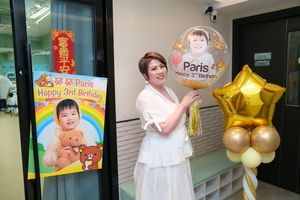 Woman in white dress poses with birthday balloons and poster celebrating Paris's 3rd birthday at a colorful party venue.