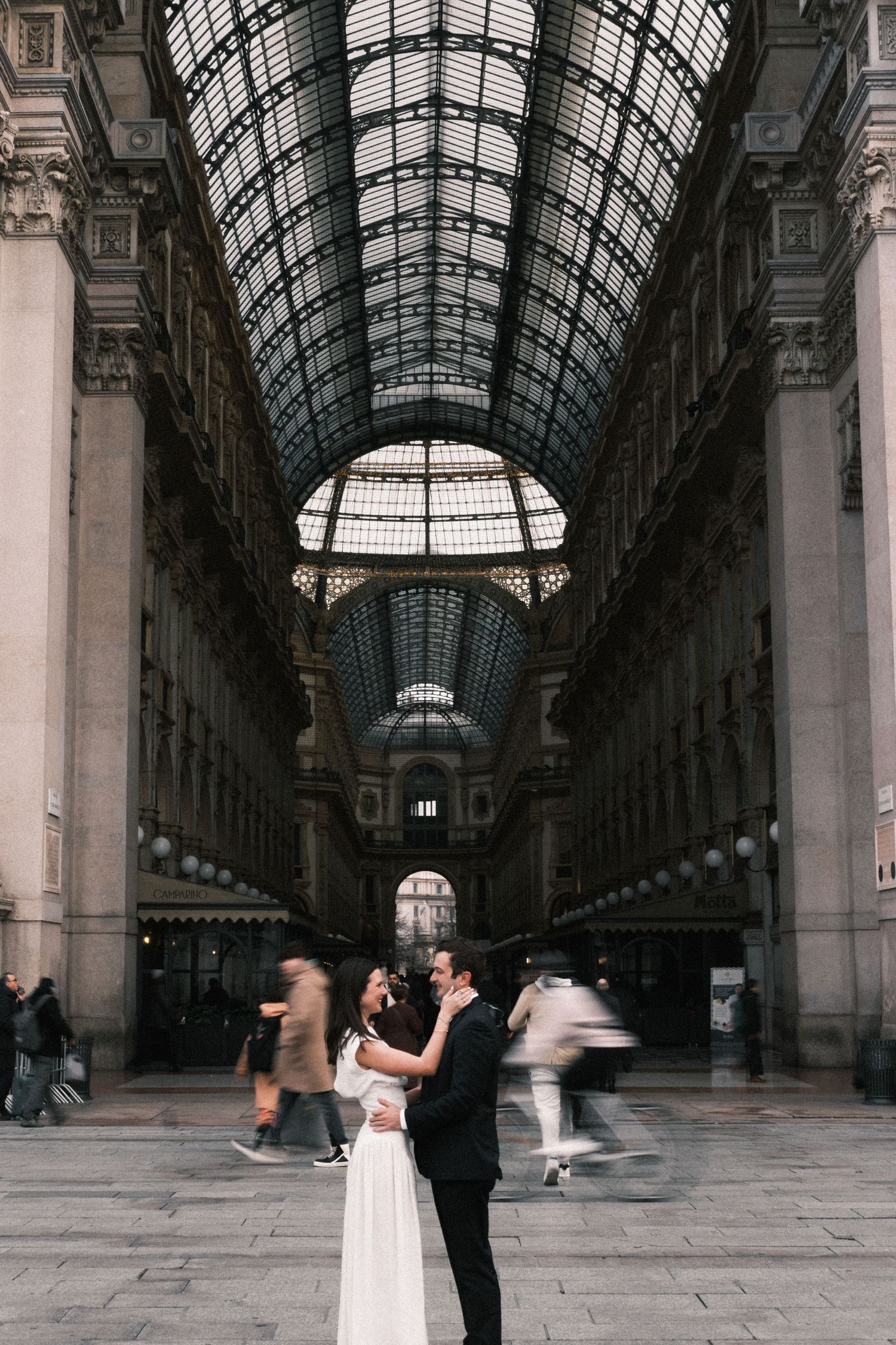 Couple embracing in Galleria Vittorio Emanuele II, Milan, with blurred pedestrians and stunning glass ceiling.