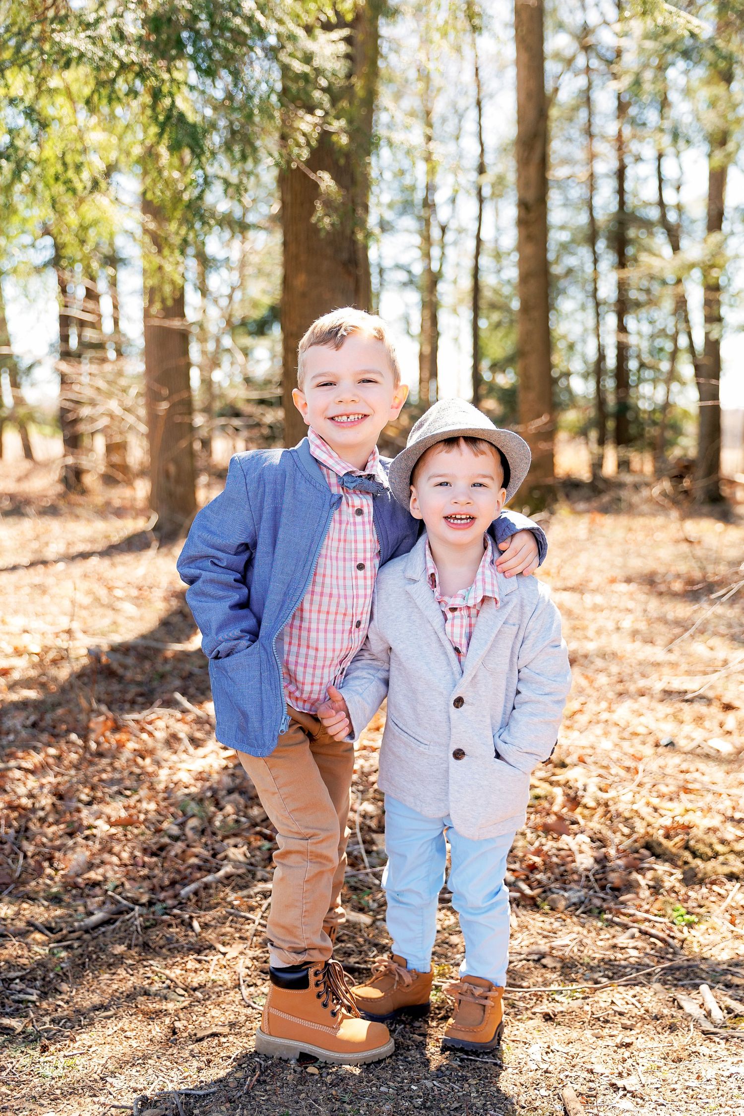 Two young boys in blazers and brown boots smile together in a sunny forest setting.