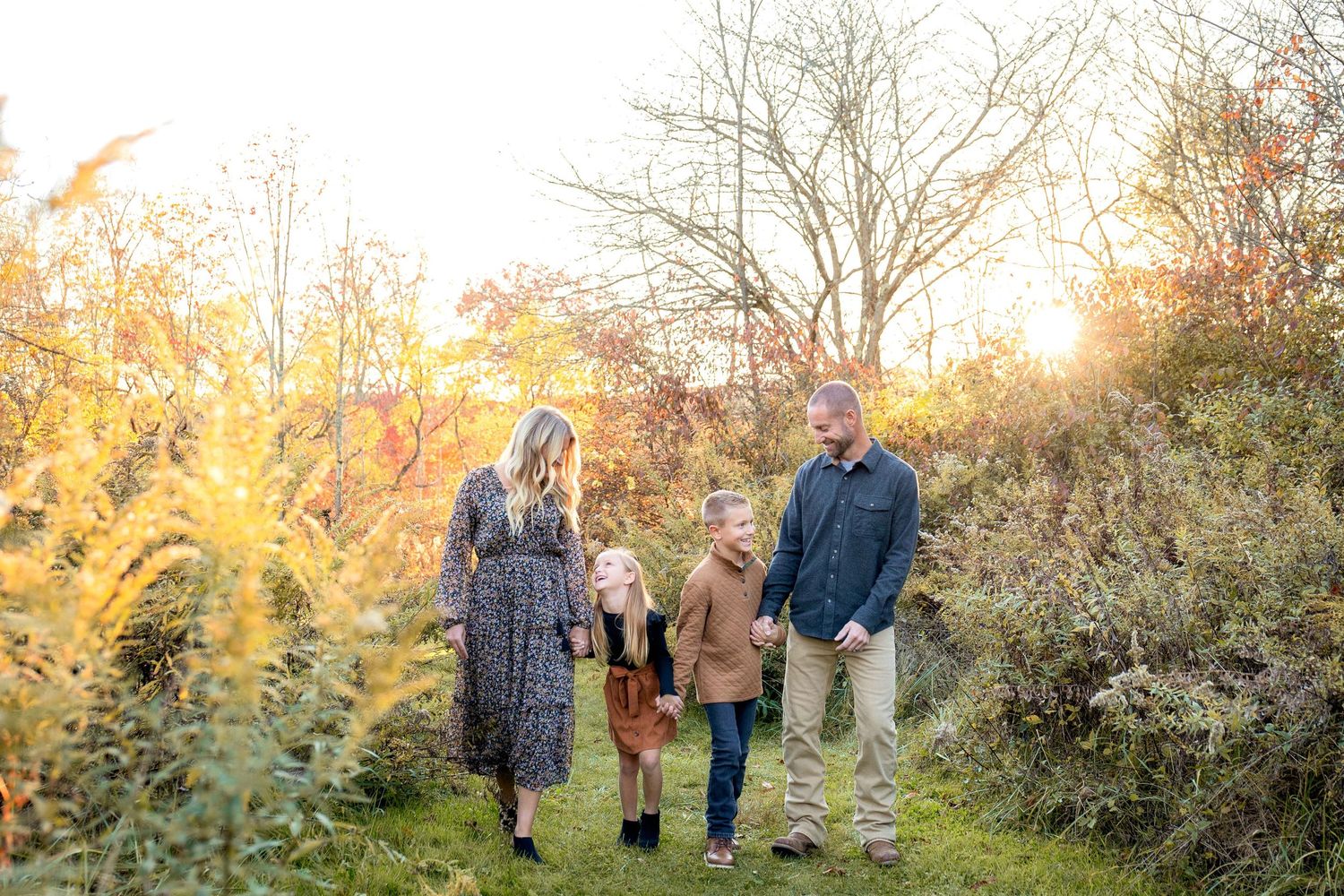 Family of four walking through golden autumn fields at sunset, parents holding hands with two children in fall foliage.
