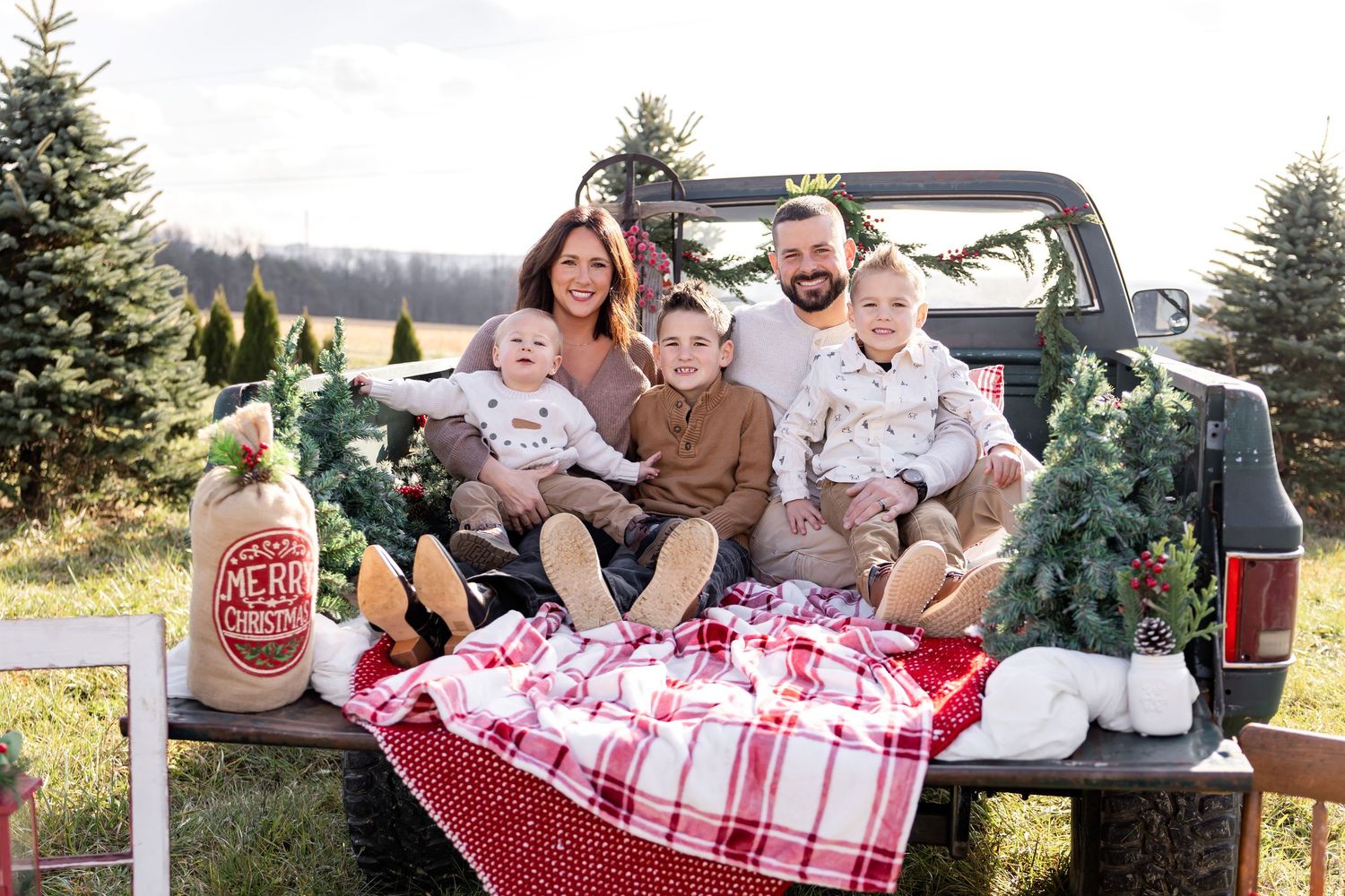 Family of five posing in truck bed with Christmas trees and holiday decor at a festive tree farm.