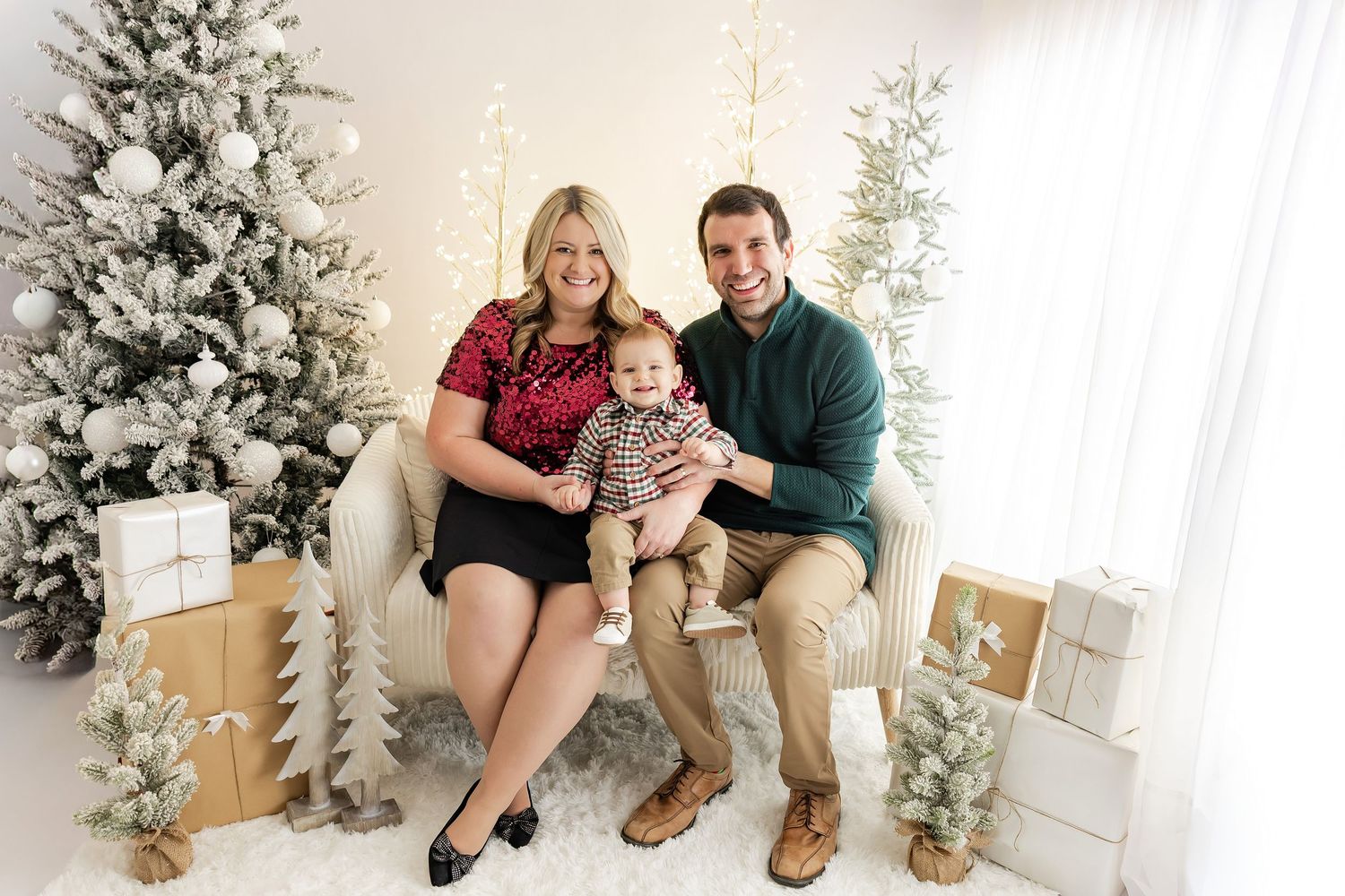 Family of three posing for a Christmas portrait with decorated trees and gifts in a bright, festive studio setting.