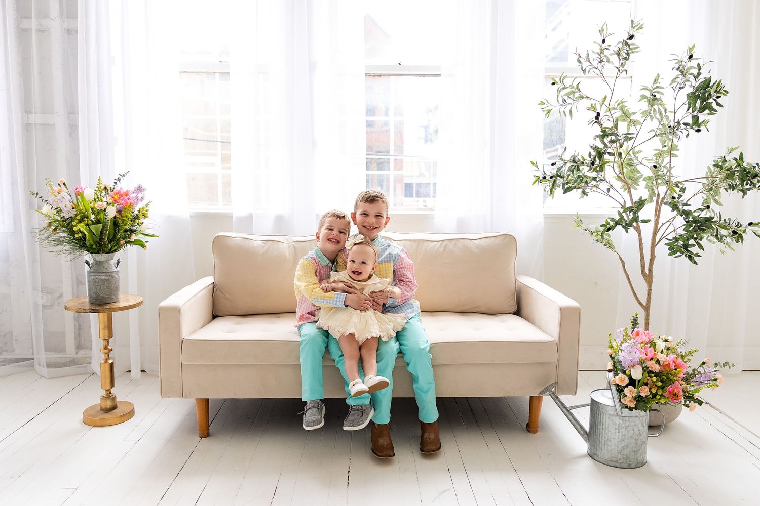 Family of three sitting together on a beige sofa in a bright white living room with flowers and plants.