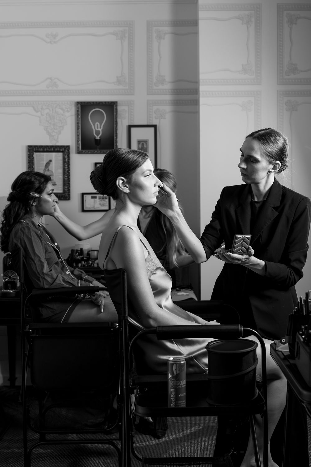 Black and white photo of a makeup artist applying cosmetics to a seated model in an elegant studio setting.