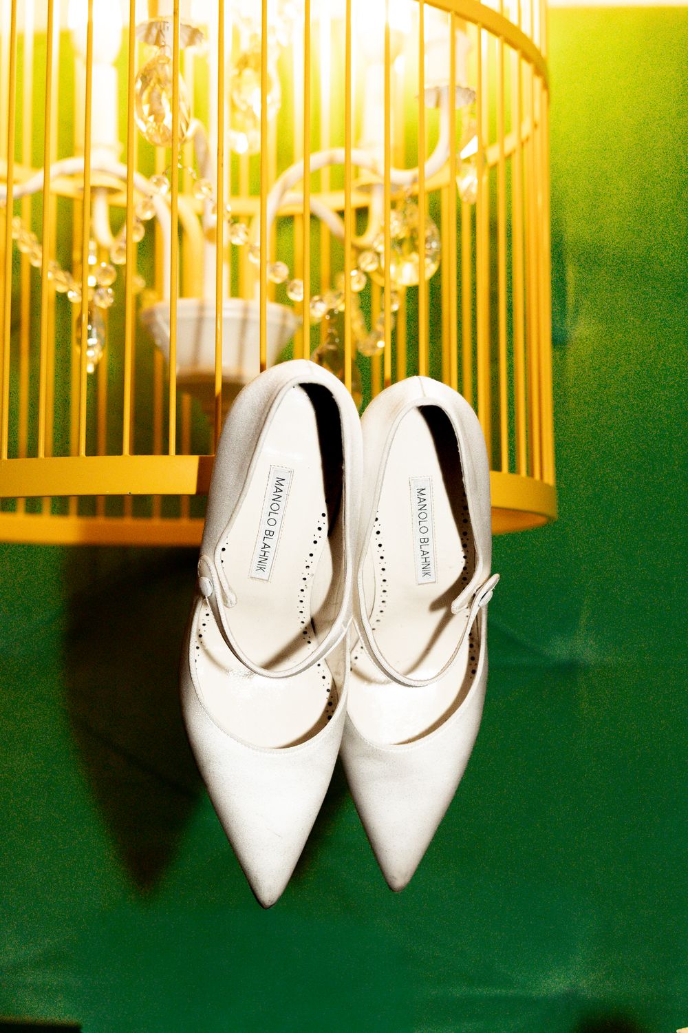 Manolo Blahnik white pointed-toe slingback heels displayed on green surface near gold birdcage decor.