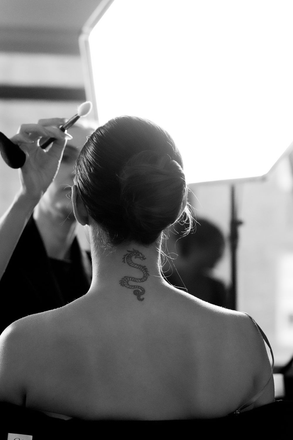 Black and white photo of woman with dragon neck tattoo getting makeup applied from behind.