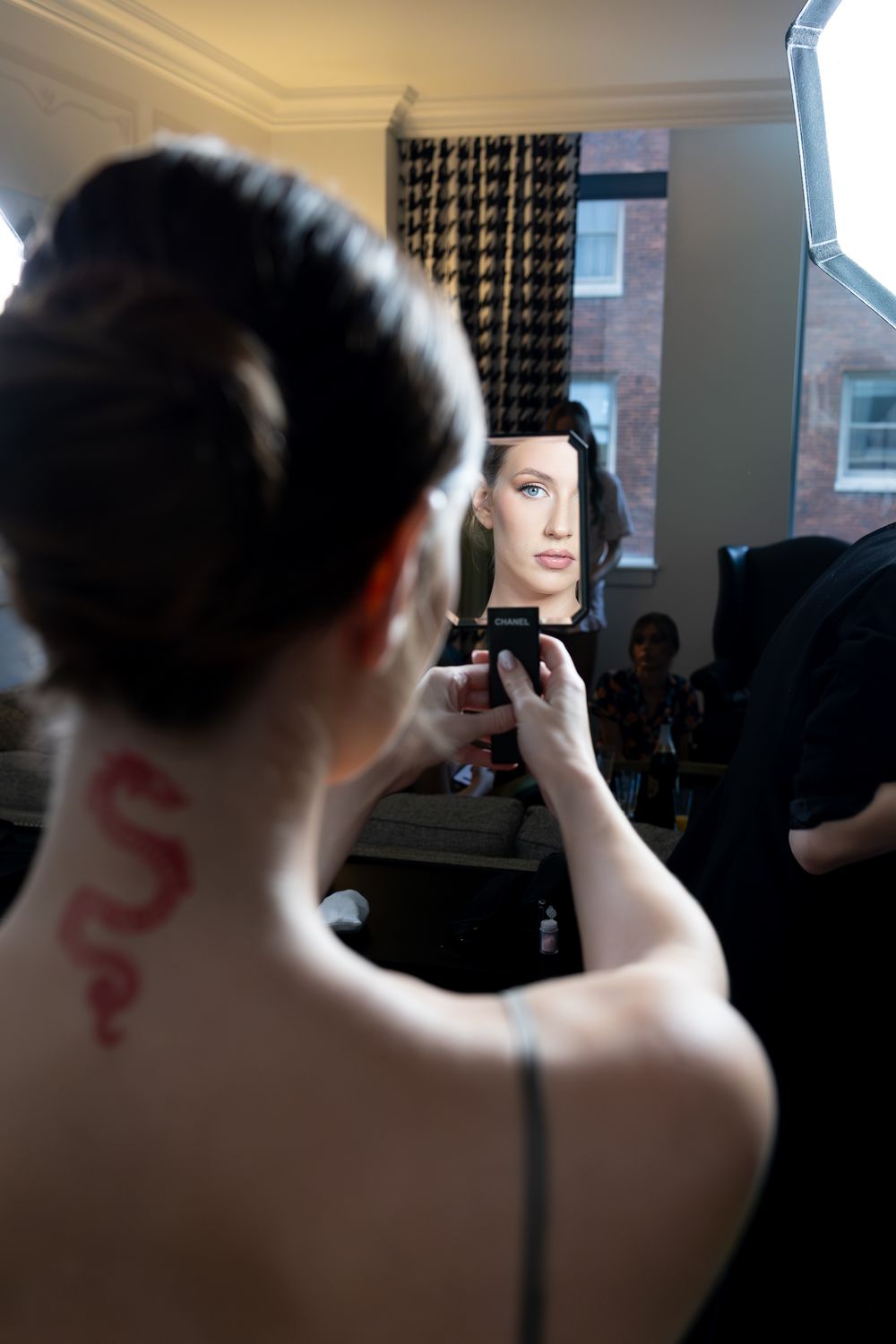 Model with snake tattoo on neck views reflection in compact mirror during backstage fashion show preparation.