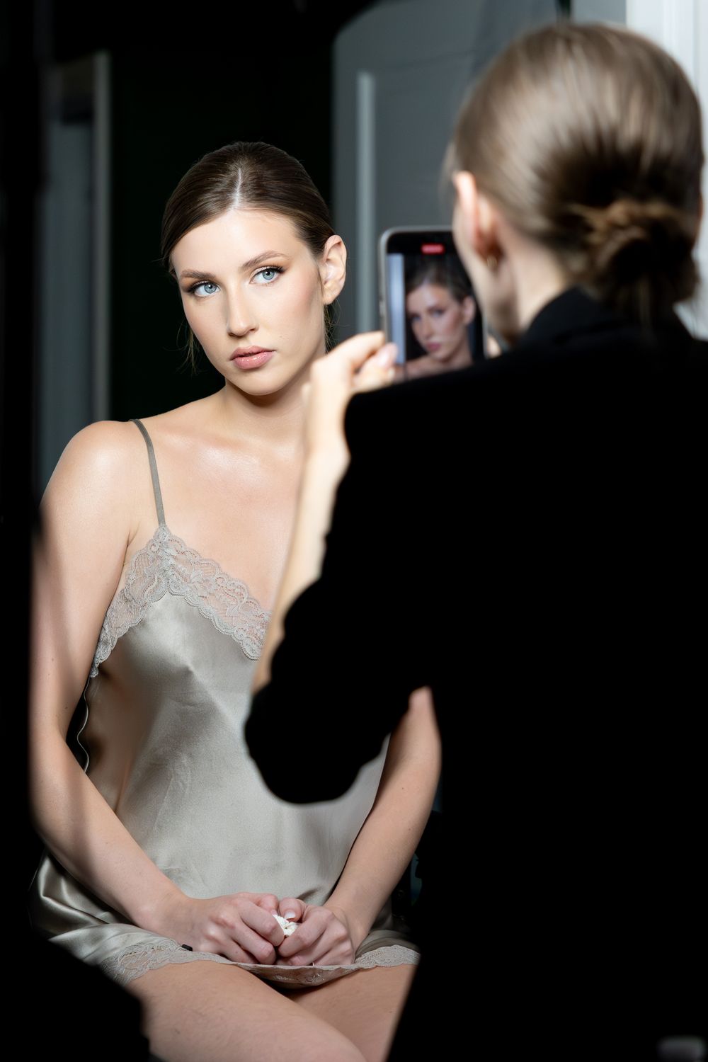 A photographer takes a photo of a young woman in a silk slip dress during a fashion backstage shoot.