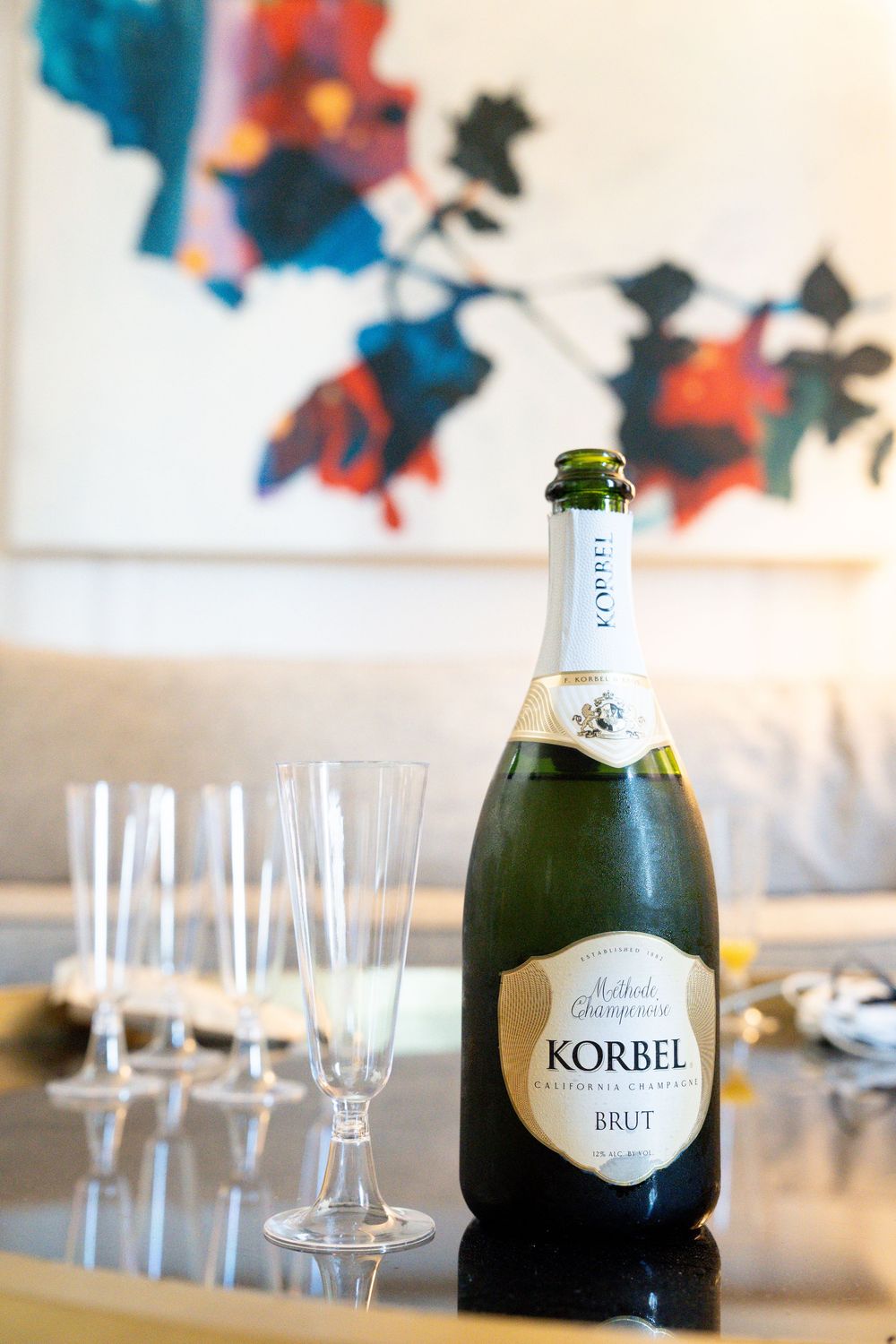 Korbel Brut Methode Champenoise sparkling wine bottle with empty champagne flutes on a table, colorful art in background.
