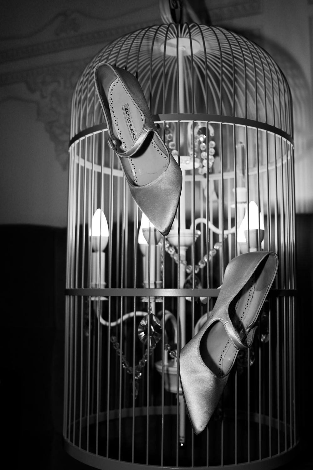 Black and white photo of elegant high heel shoes hanging inside a decorative birdcage with chandelier lighting.