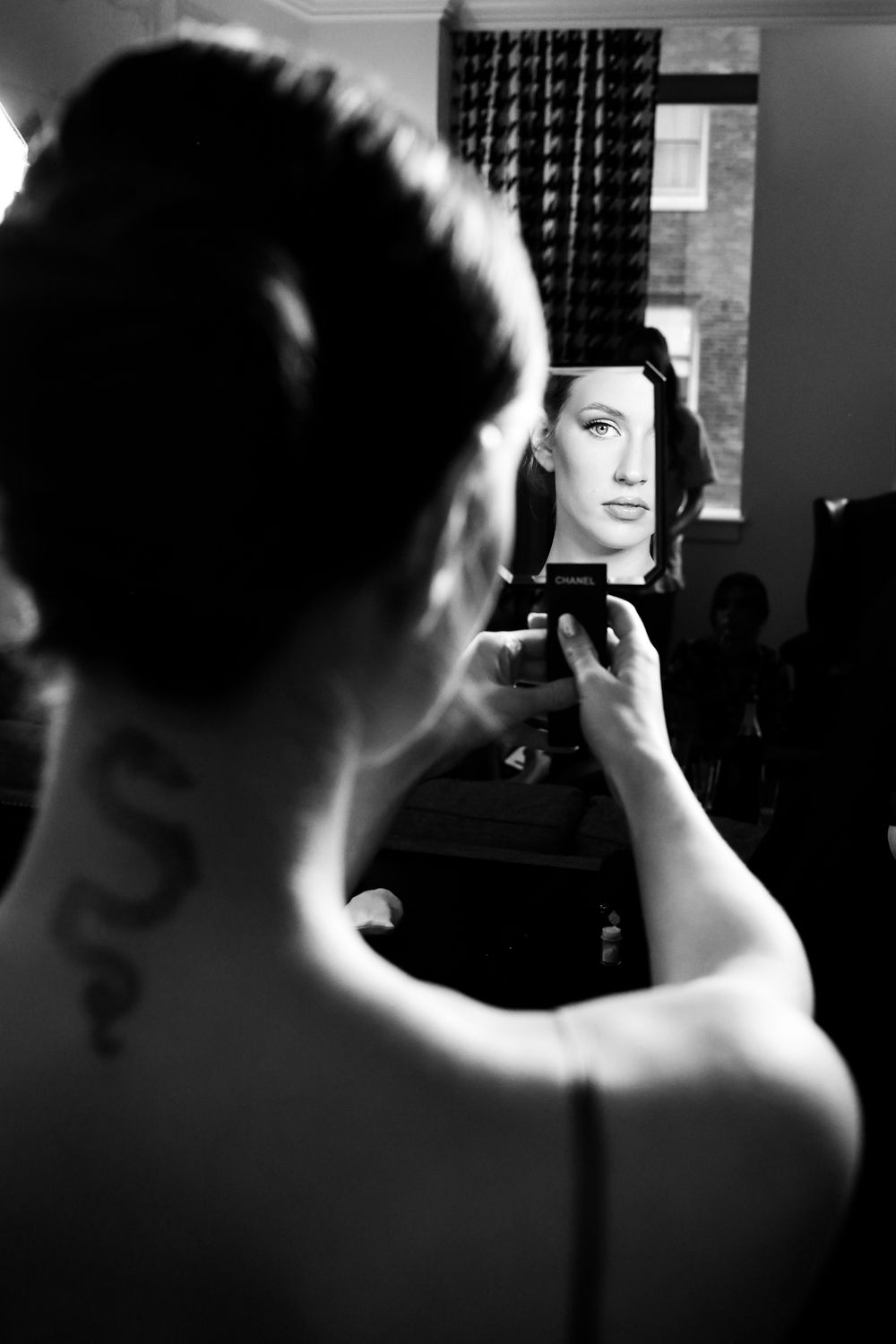 Black and white photo of woman with neck tattoo holding small mirror reflecting her face.
