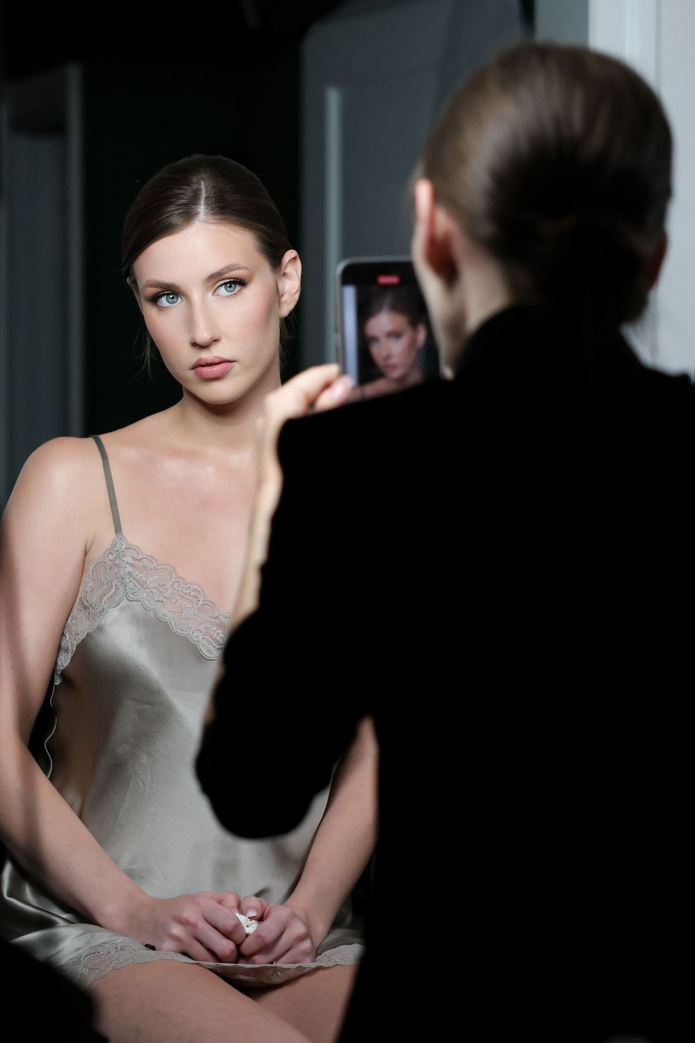 Photographer taking a photo of an elegant young woman in a silk slip dress using a smartphone in a studio.