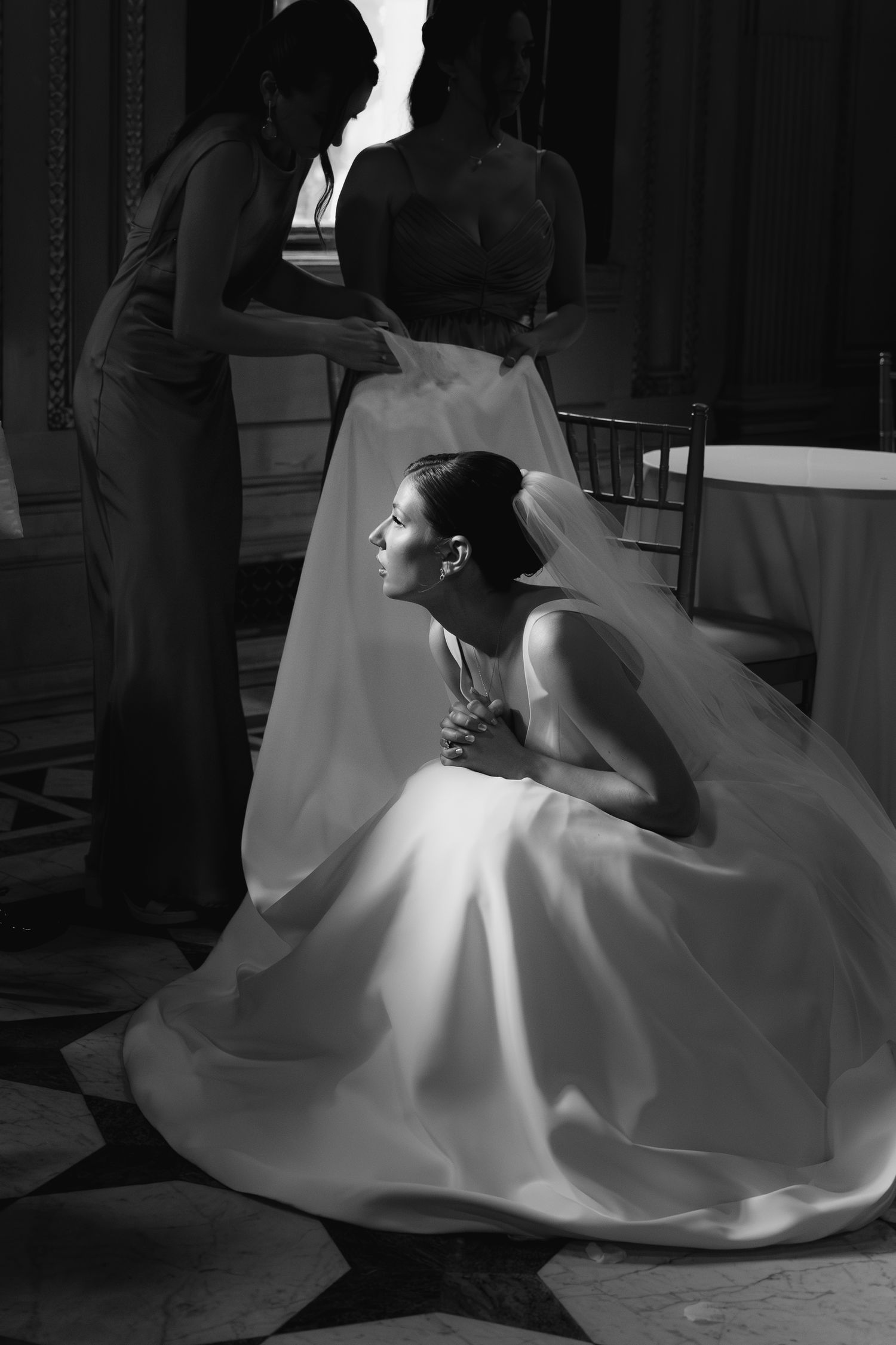 Black and white photo of a bride in a flowing gown kneeling gracefully while bridesmaids adjust her veil.