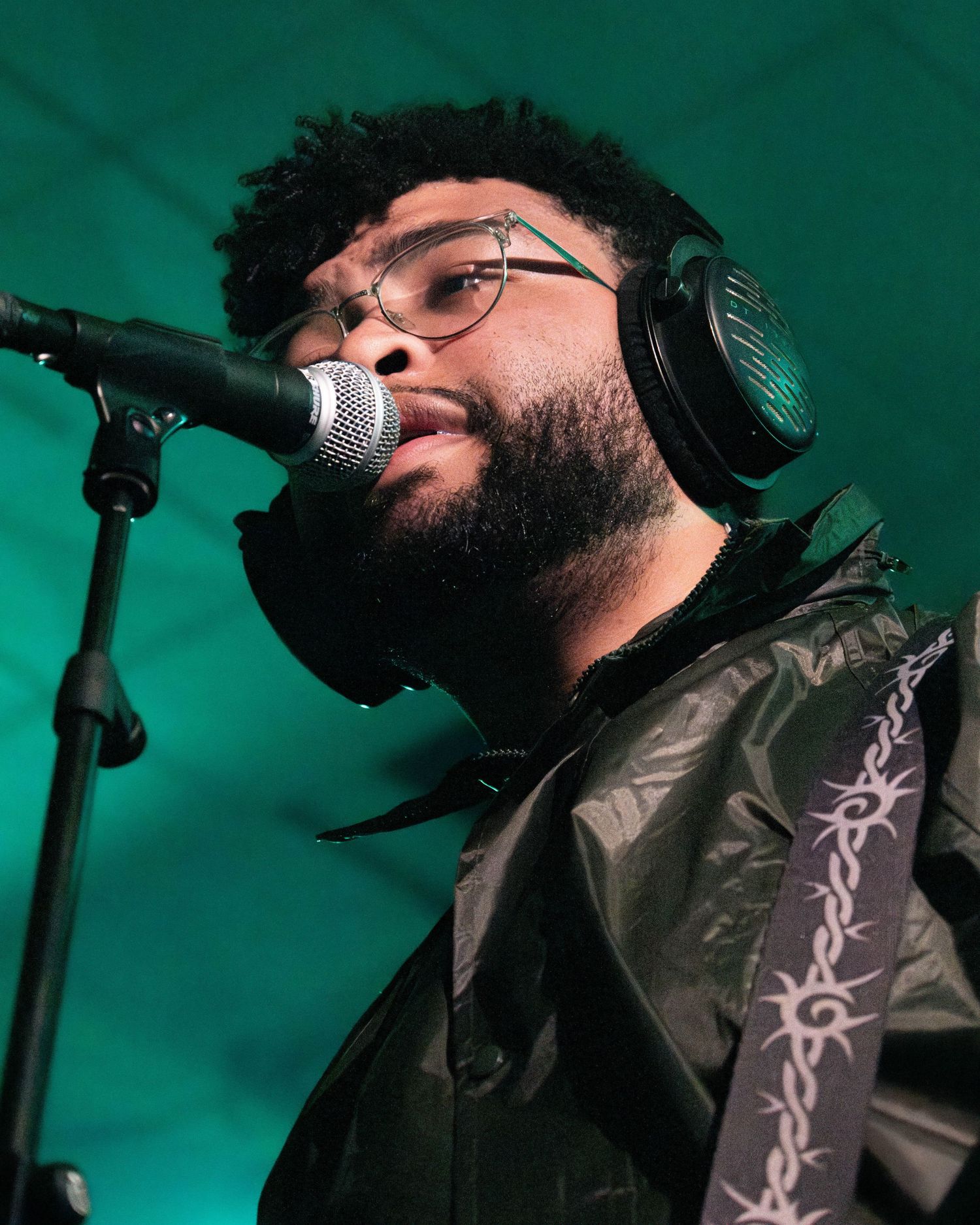 Young man with headphones and glasses singing into a microphone on stage wearing a dark jacket.