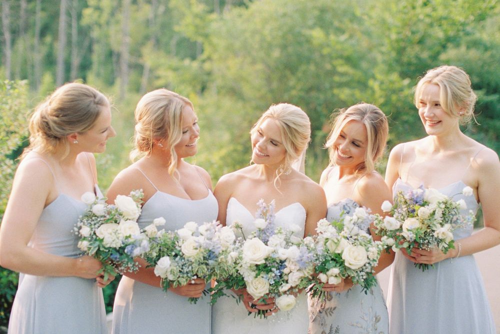 Bride and four bridesmaids in light gray gowns holding white floral bouquets, smiling outdoors with lush greenery background.