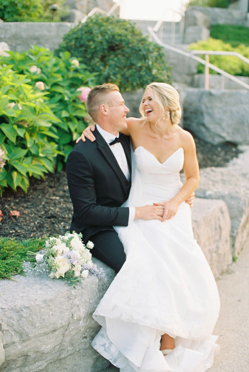 A laughing bride in a strapless white gown sits with her tuxedo-clad groom on stone steps surrounded by lush greenery.