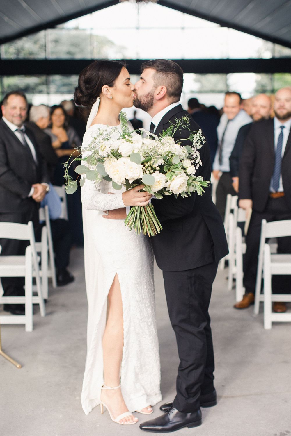 Bride and groom share first kiss at wedding ceremony, bride holding white floral bouquet, guests seated in white chairs.