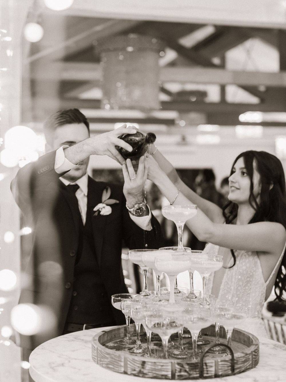 Bride and groom pouring champagne tower at wedding reception in elegant black and white photo.