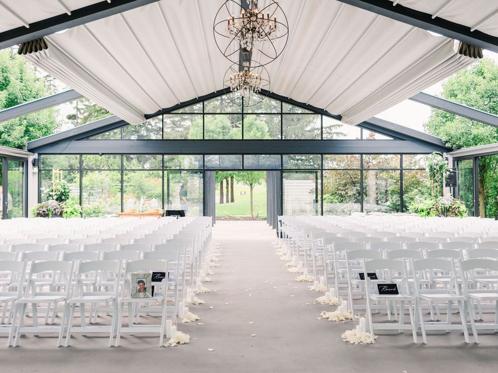 Elegant wedding ceremony venue with white chairs, floral aisle decor, glass walls, and a chandelier under a vaulted ceiling.