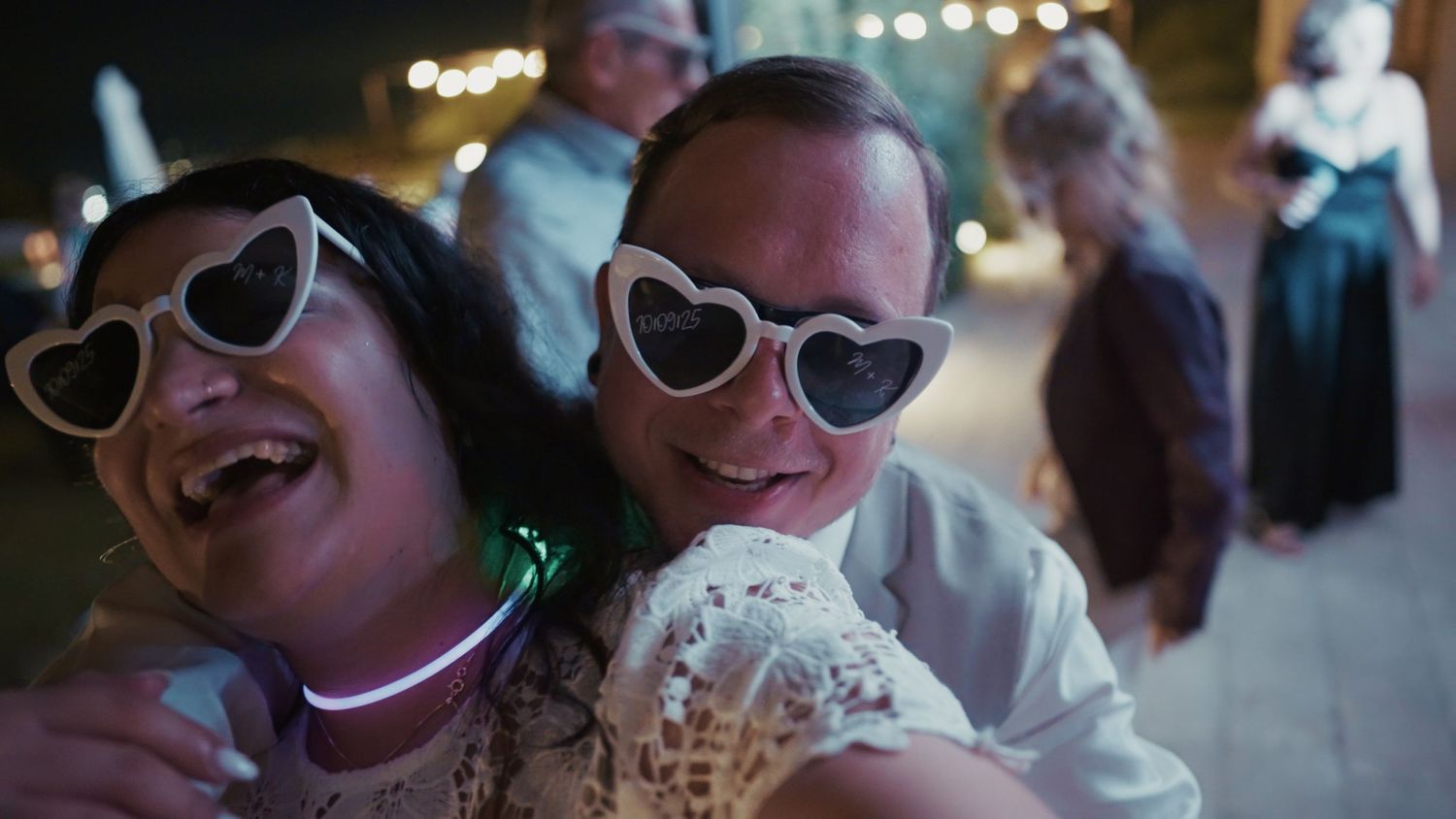 Two people wearing heart-shaped sunglasses share a joyful moment at an outdoor wedding reception at night.