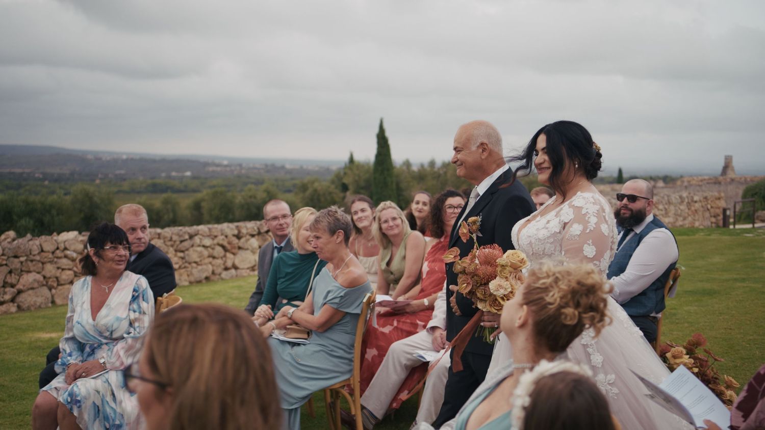 Outdoor wedding ceremony with bride and groom surrounded by guests, stone wall and green landscape in background.