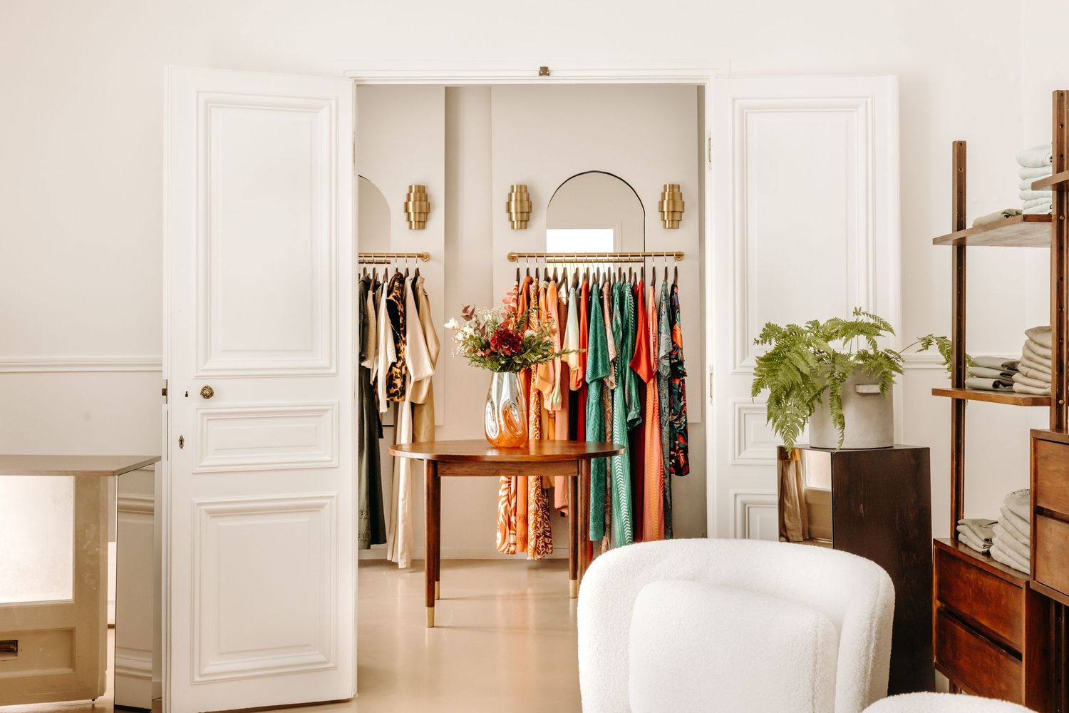 Elegant boutique dressing room with white French doors, colorful clothing racks, gold wall sconces, and lush greenery accents.
