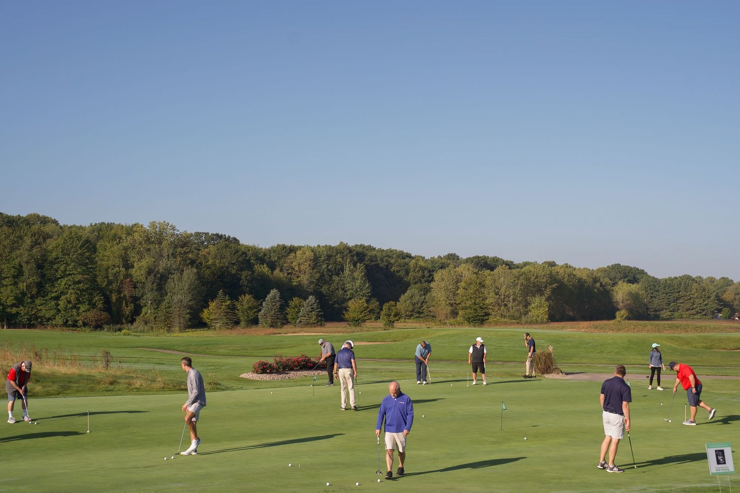 Golfers practice on a sunny green fairway surrounded by autumn trees at a golf course.