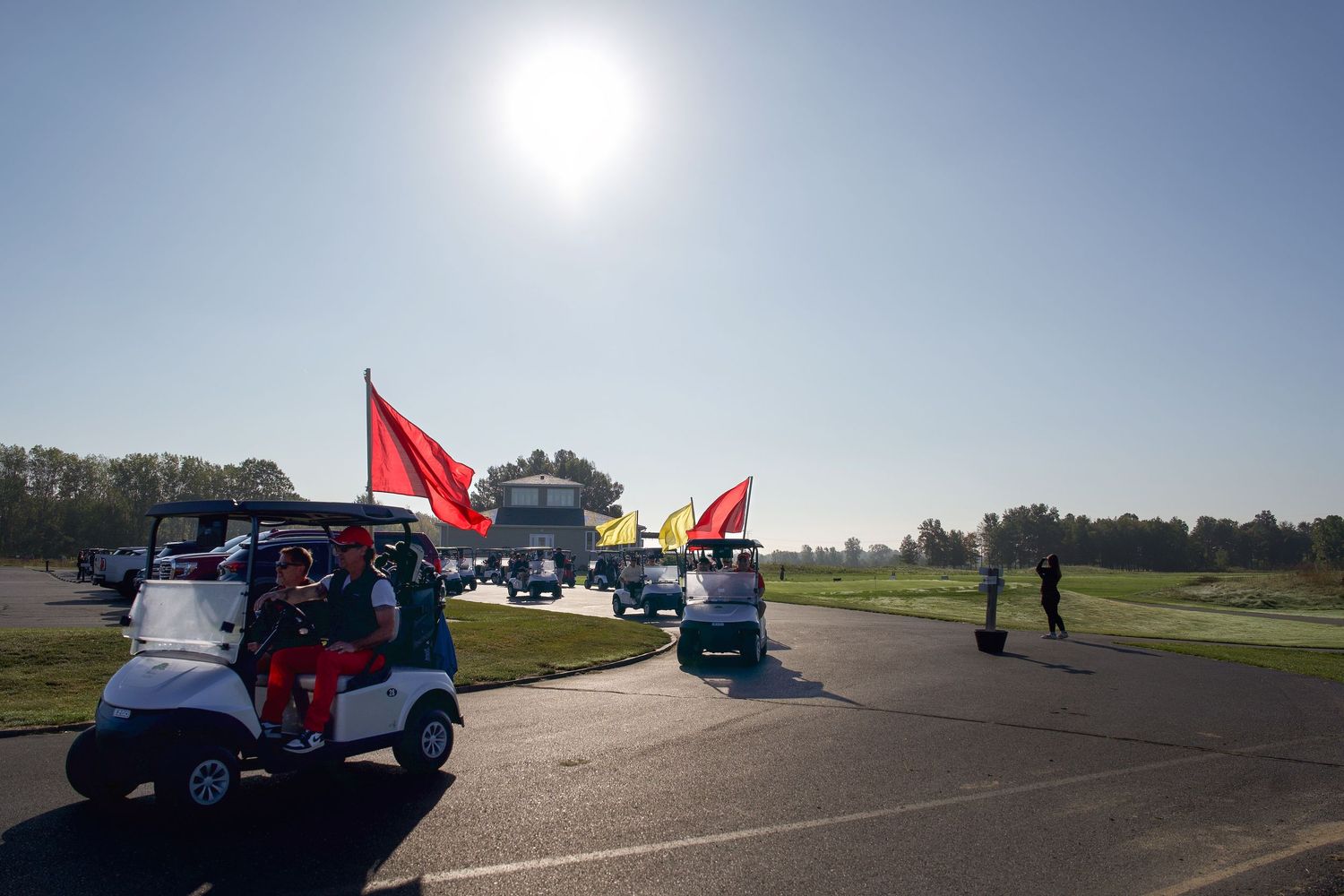 Golf carts decorated with red flags line up on a sunny golf course path during an outdoor event.