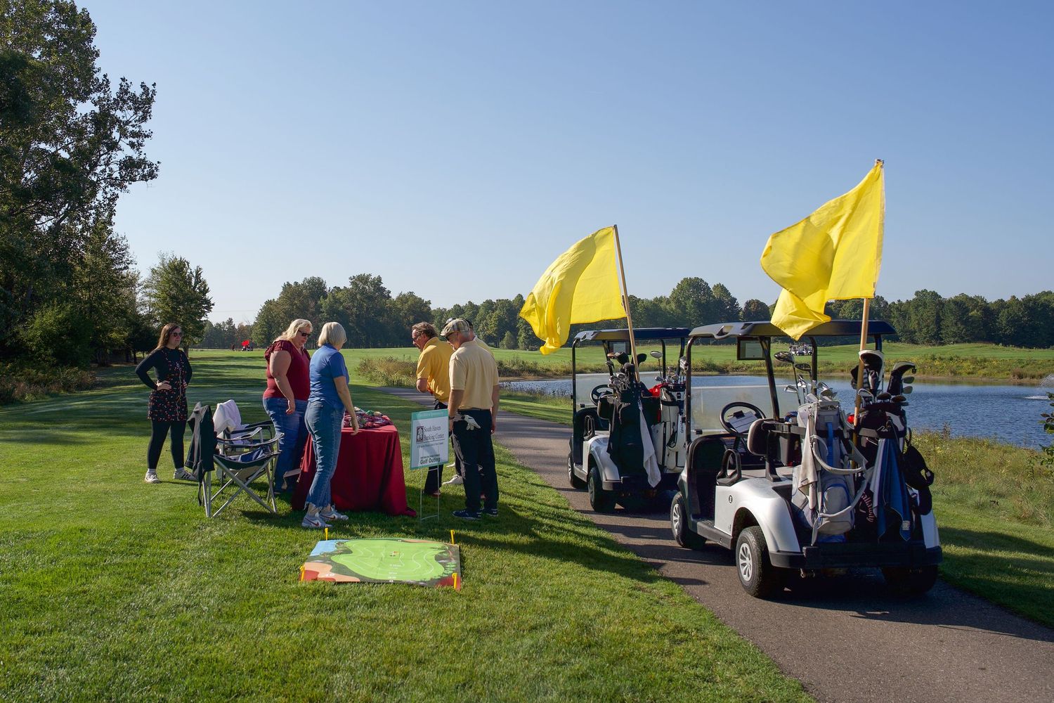 Golfers gather near yellow-flagged golf carts on a sunny green course with a pond and trees in the background.