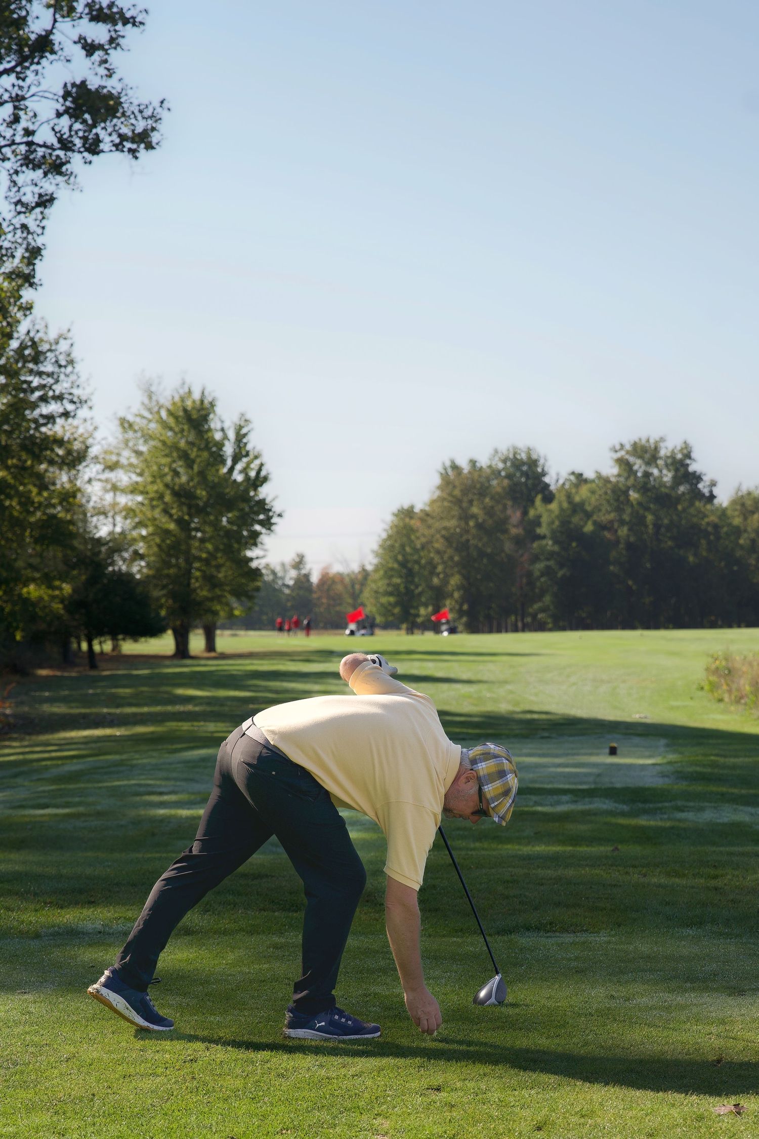 Golfer bending down to place a tee on a sunny green golf course with trees in the background.