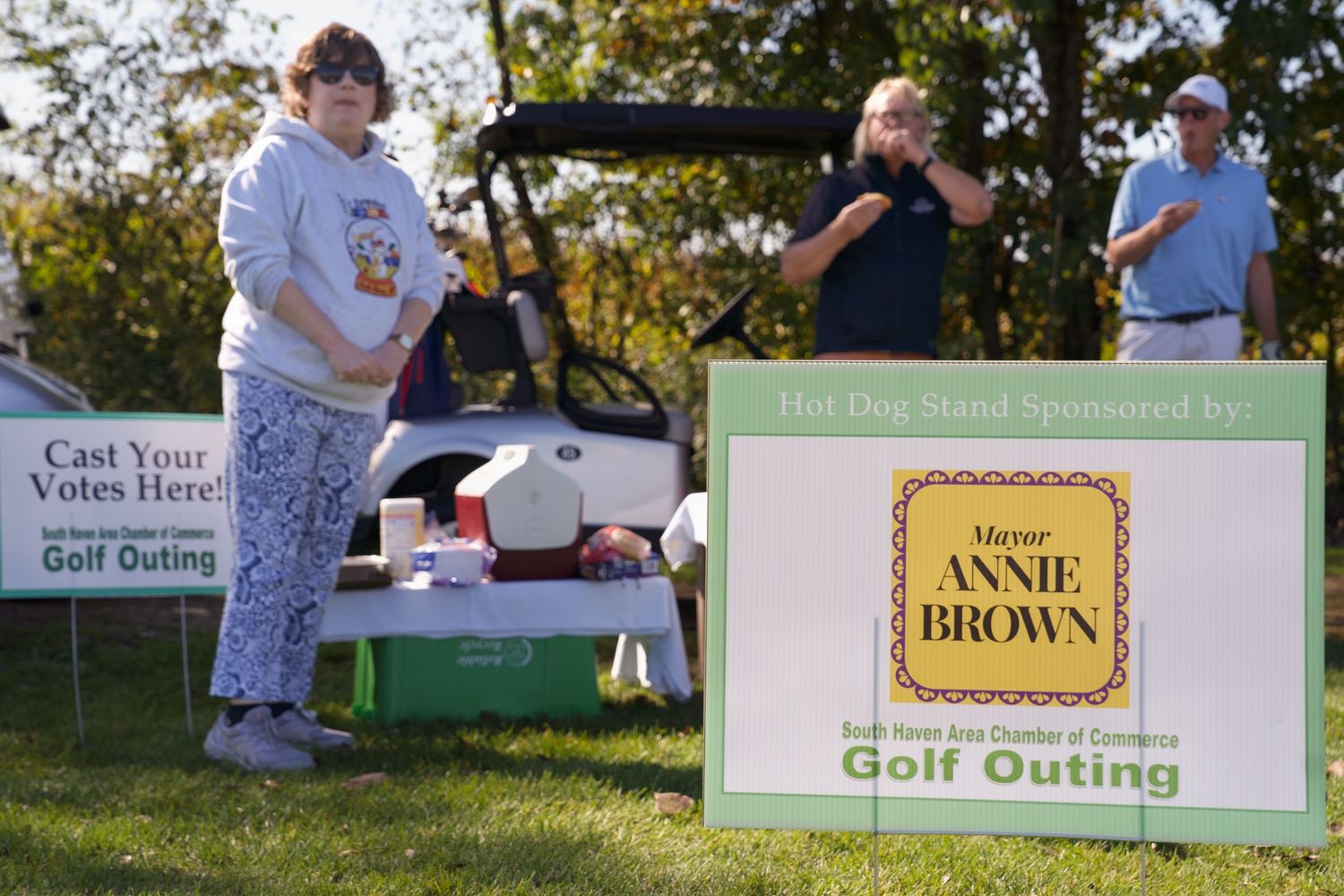 Annie Brown Mayor Golf Outing sign at hot dog stand sponsored event with attendees and golf cart on sunny day.