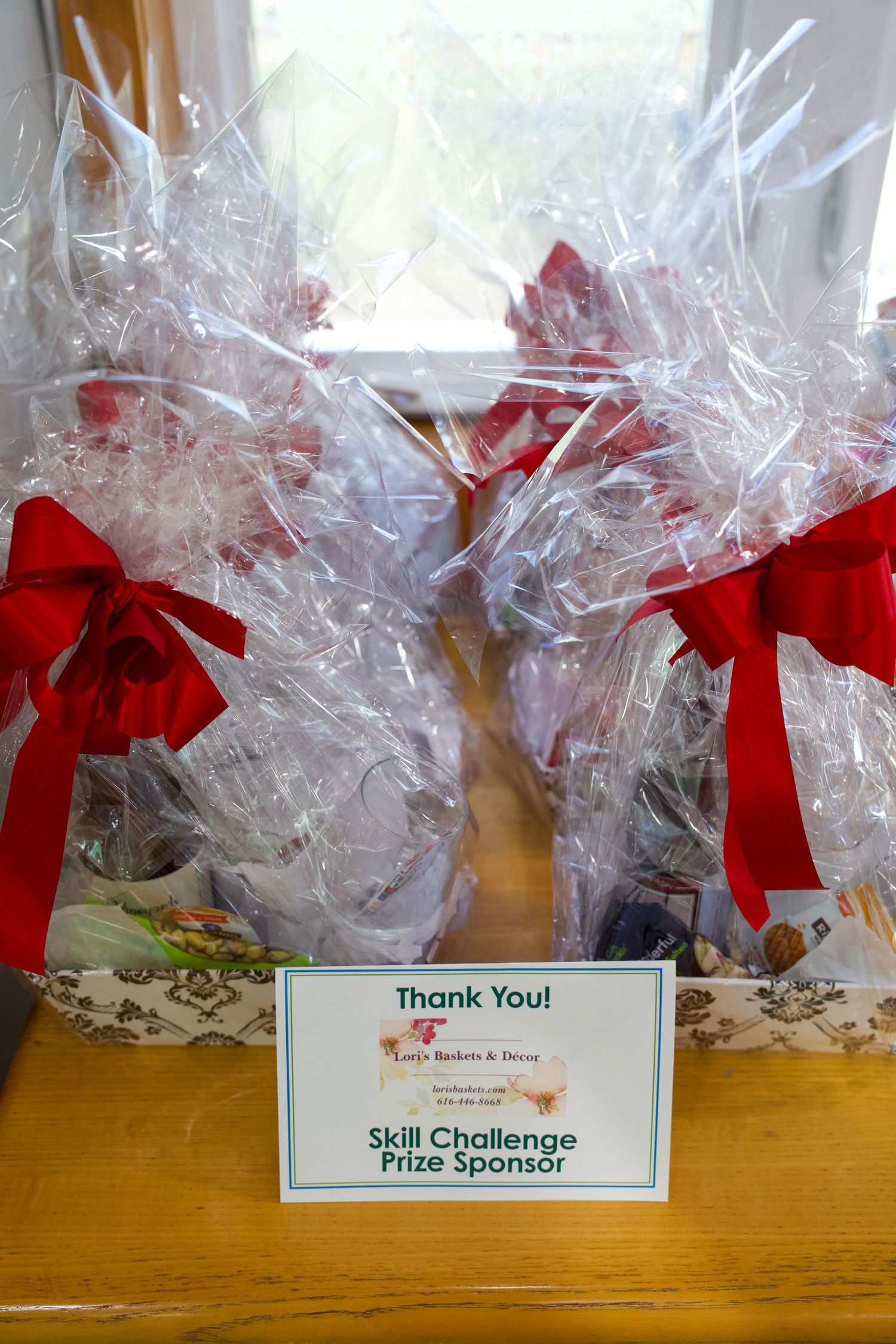 Two gift baskets wrapped in clear cellophane with red bows and a Thank You Skill Challenge Prize Sponsor sign on a wooden table.