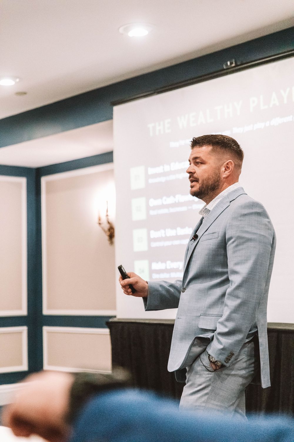 A man in a gray suit presents The Wealthy Play slideshow to an audience in a conference room.