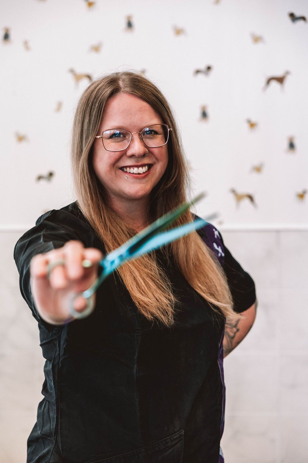 Smiling woman with glasses holding teal scissors, standing in front of a white wall decorated with small dog silhouettes.
