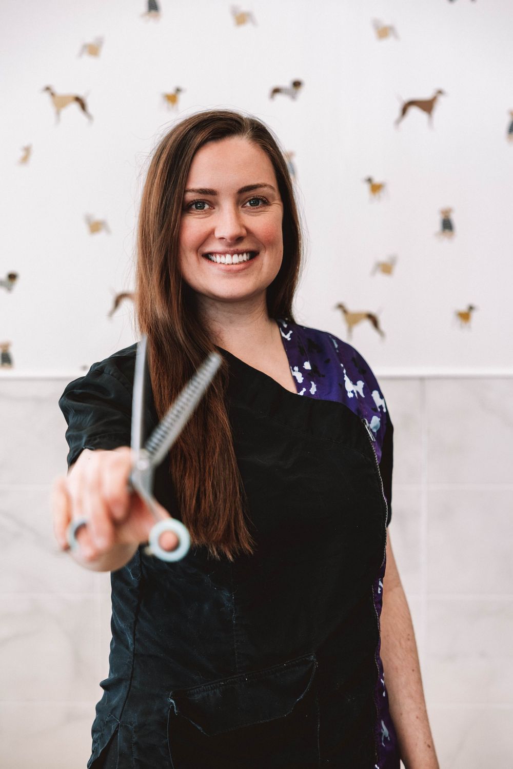 Smiling female dog groomer holding thinning scissors in a salon with dog illustrations on the wall.