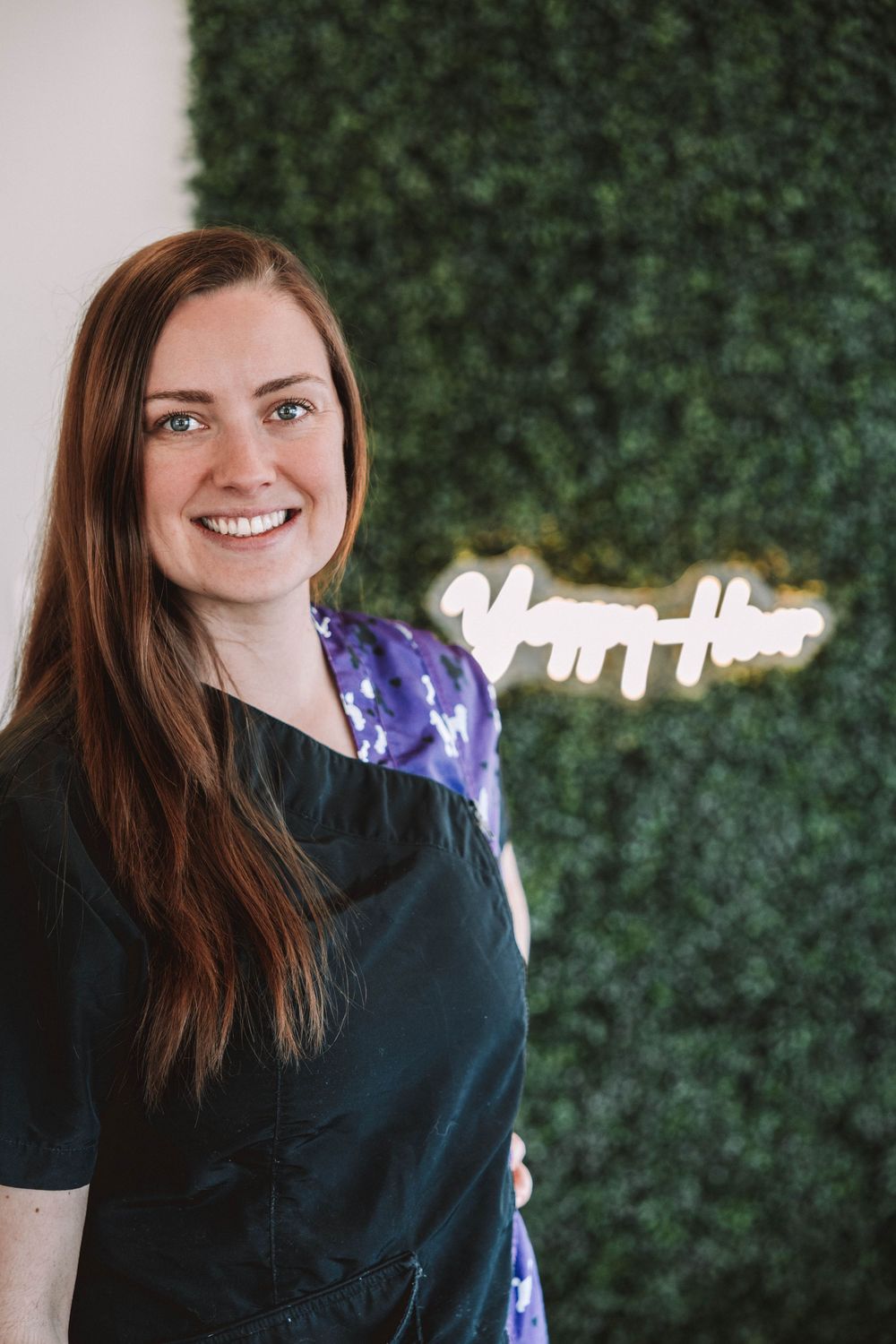 Smiling woman with long red hair wearing a dark apron stands before a green hedge wall with a neon sign.