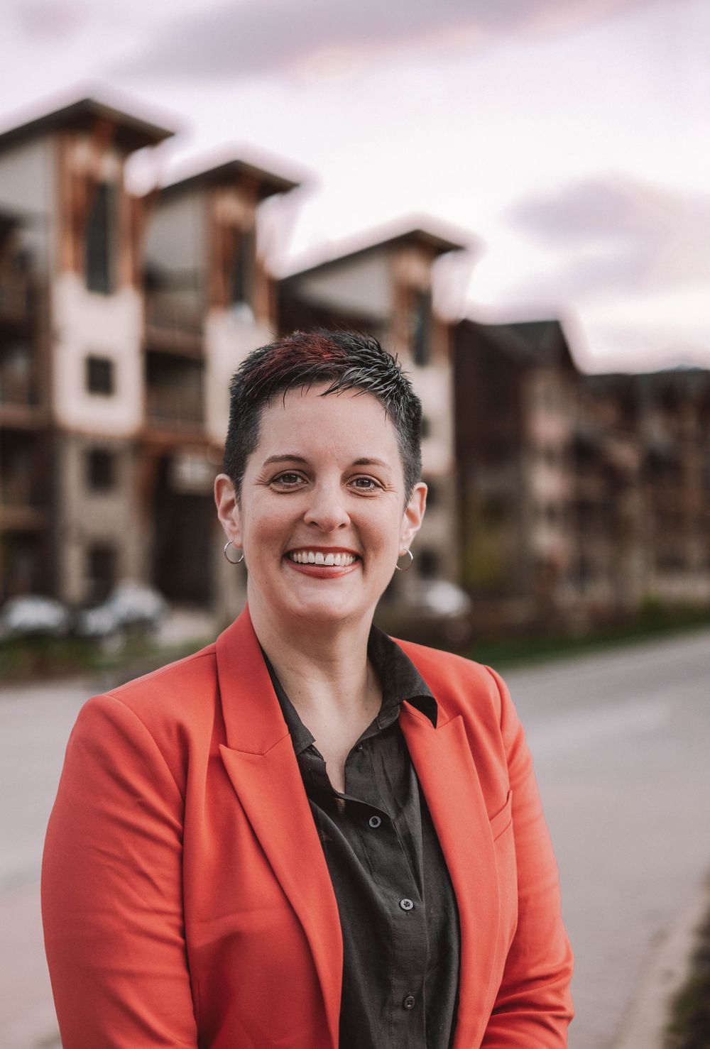 A smiling woman in a red blazer stands outdoors near modern apartment buildings at dusk.