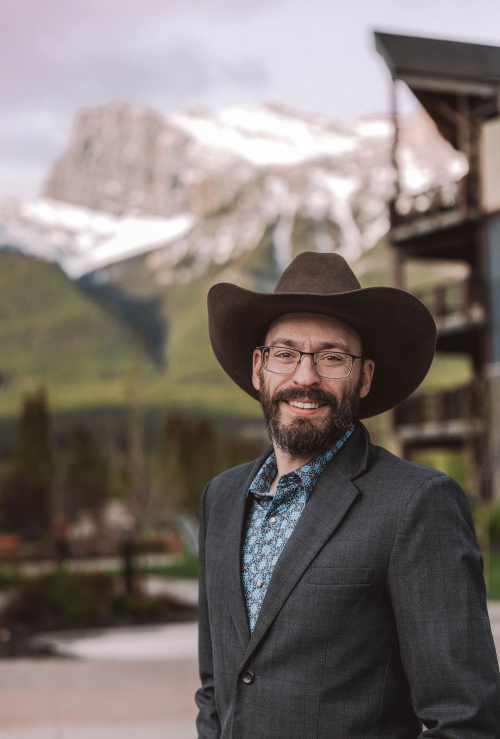 Smiling bearded man in cowboy hat, glasses, and blazer posing outdoors with snow-capped mountains in background.