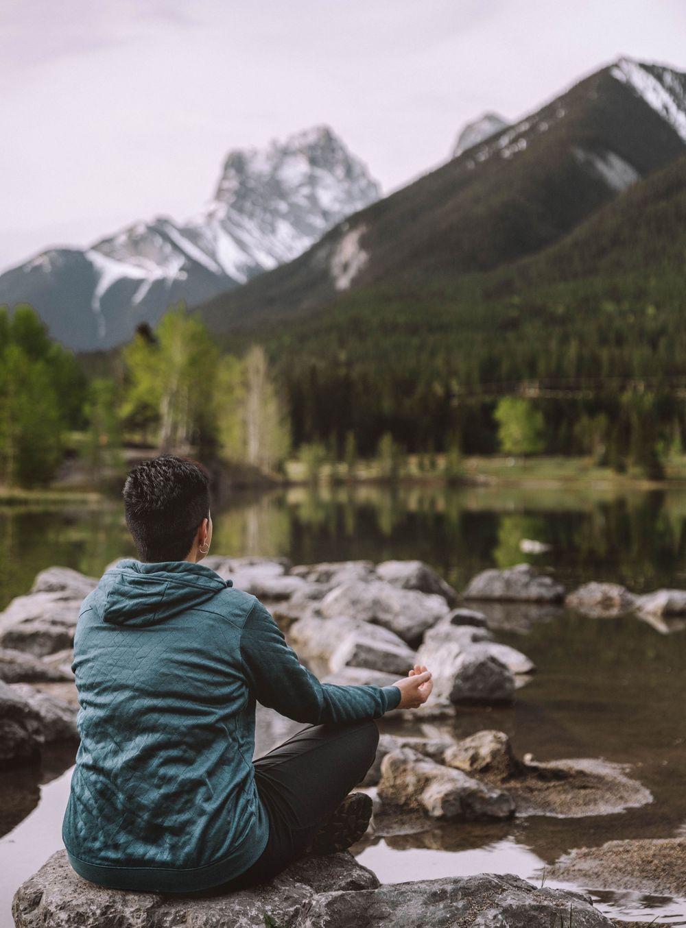 A man sits cross-legged on rocks by a calm mountain lake, gazing at snow-capped peaks reflected in still water.