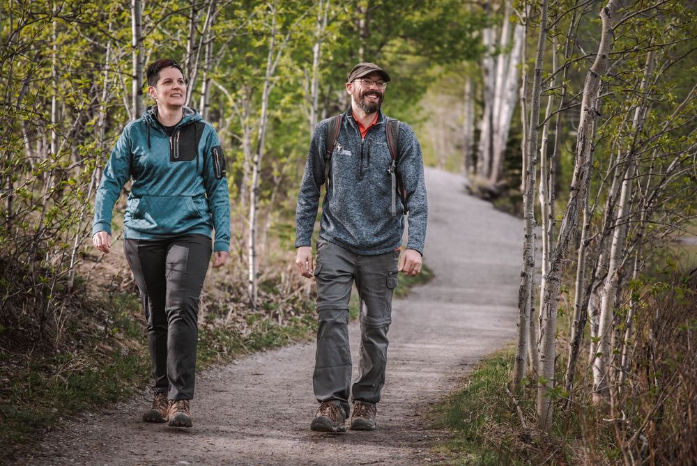 Two hikers walking on a forest trail surrounded by birch trees, wearing backpacks and outdoor clothing.