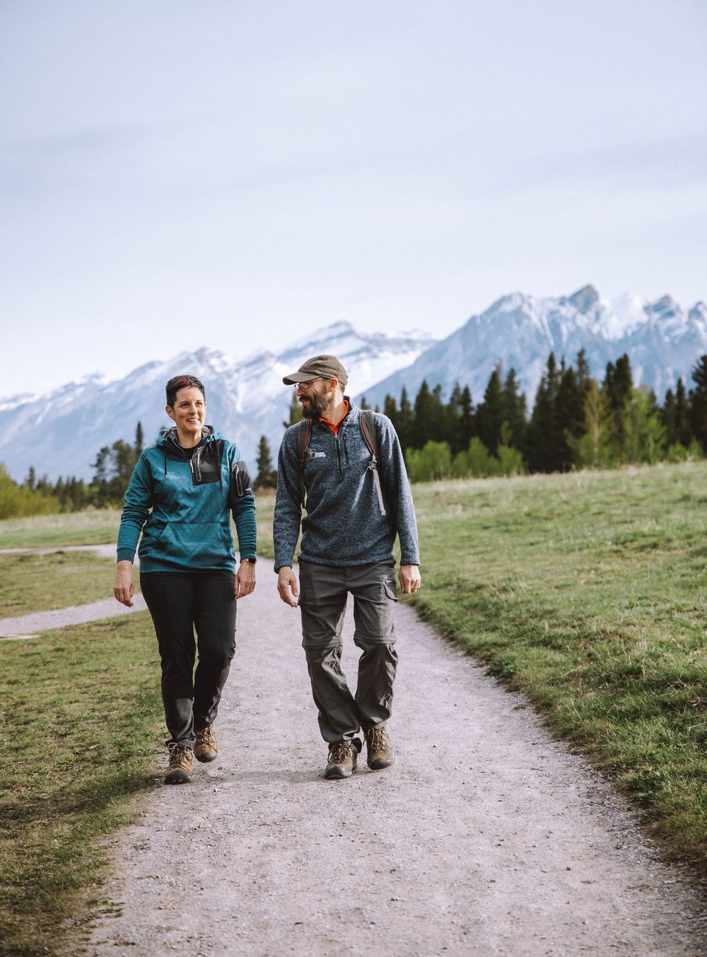 Couple hiking on a mountain trail with snow-capped peaks and green meadows in the background.