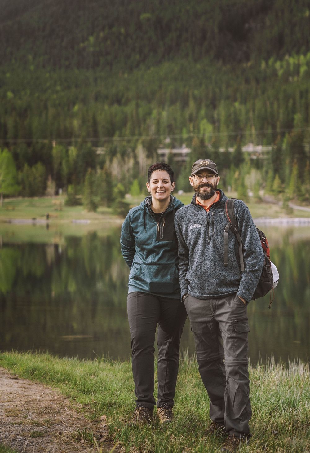 A couple smiles together near a mountain lake surrounded by evergreen forest on a hiking trail.