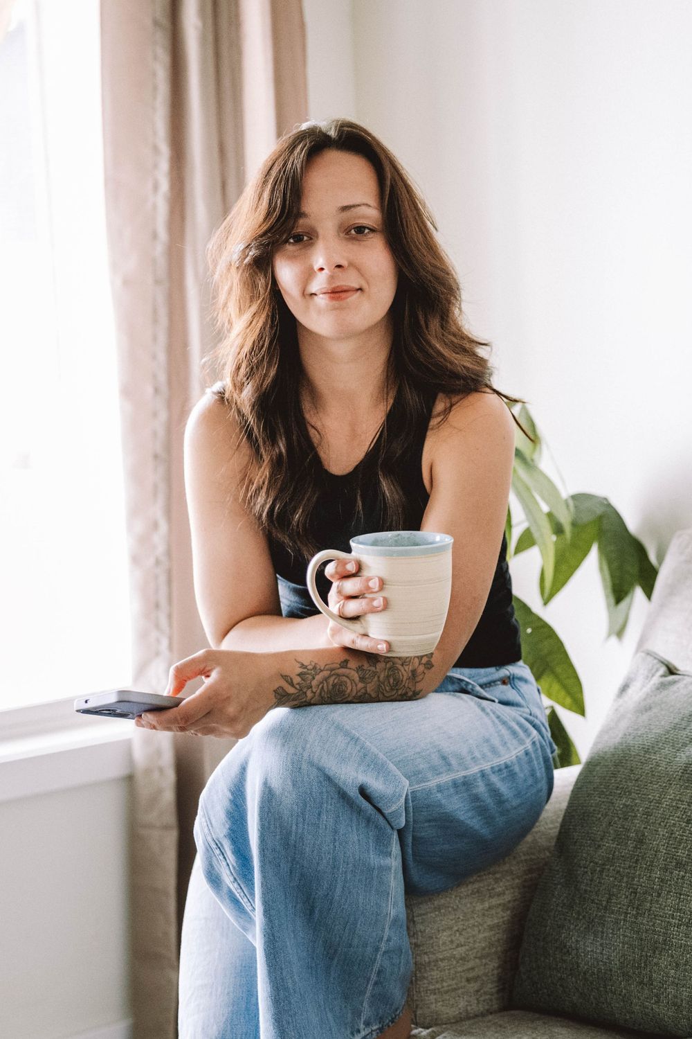 Woman with tattoo sits on couch holding coffee mug and phone, smiling near bright window with plant.
