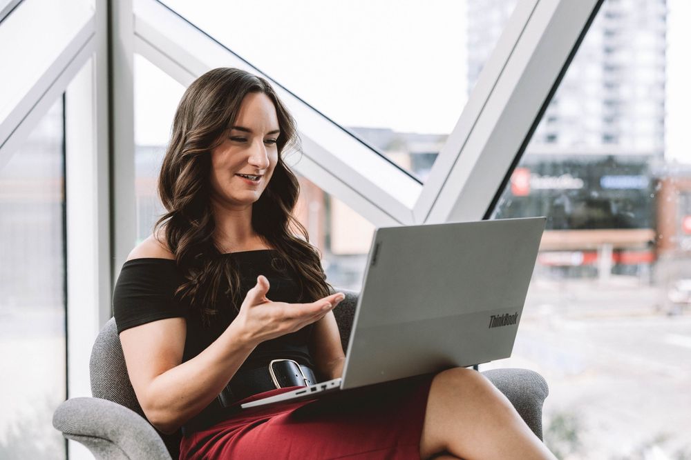 Smiling woman with long brown hair gestures during a video call on her laptop while seated in a chair by large city-view windows.