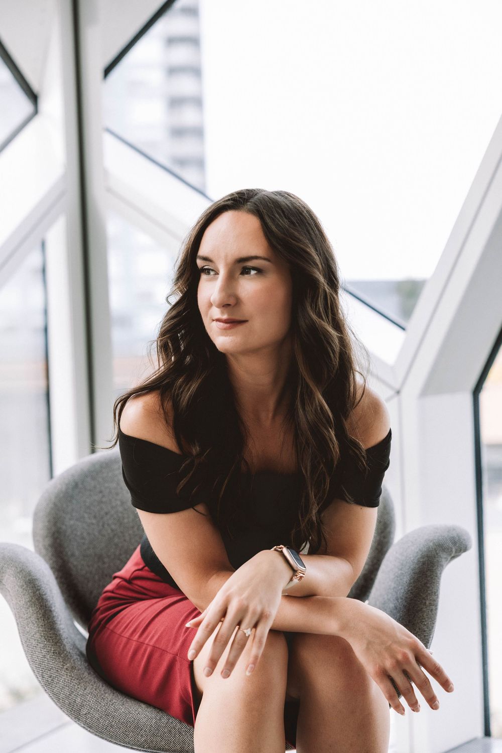 Woman with long wavy brown hair in an off-shoulder black top and red skirt sits in a gray chair beside large windows, gazing away.