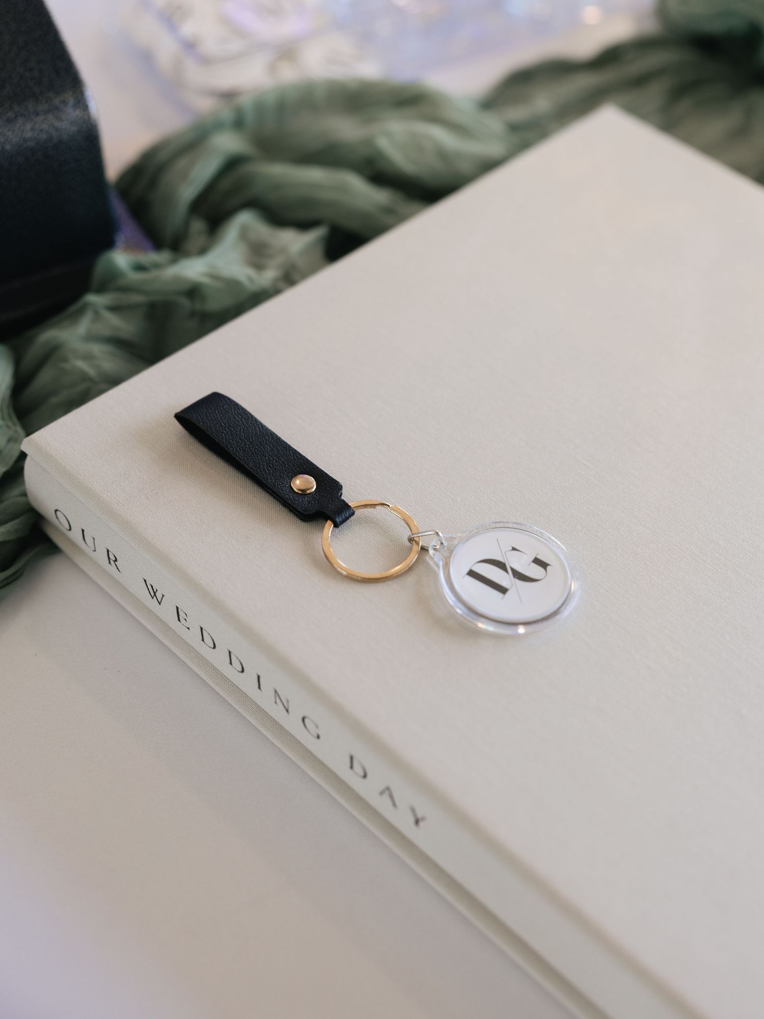 A gold keychain with acrylic monogram pendant and black leather strap resting on a white Our Wedding Day book.