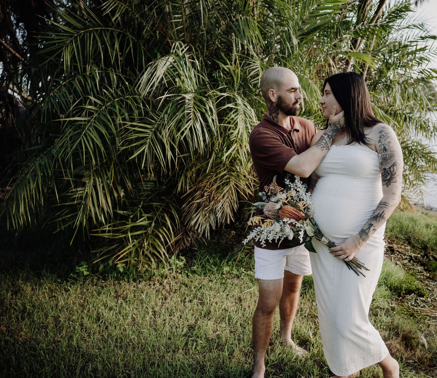 Pregnant couple embracing outdoors near tropical palm trees, woman in white dress with tattoos holding a bouquet of flowers.