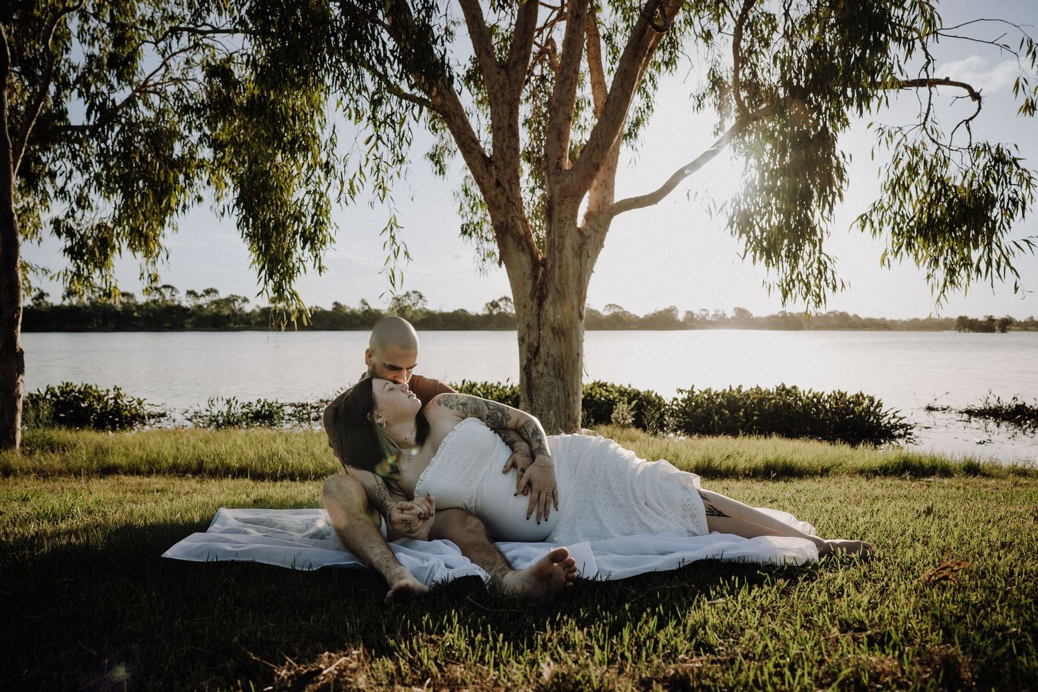 Pregnant couple embracing outdoors near tropical palm trees, woman in white dress with tattoos holding a bouquet of flowers. maternity photo