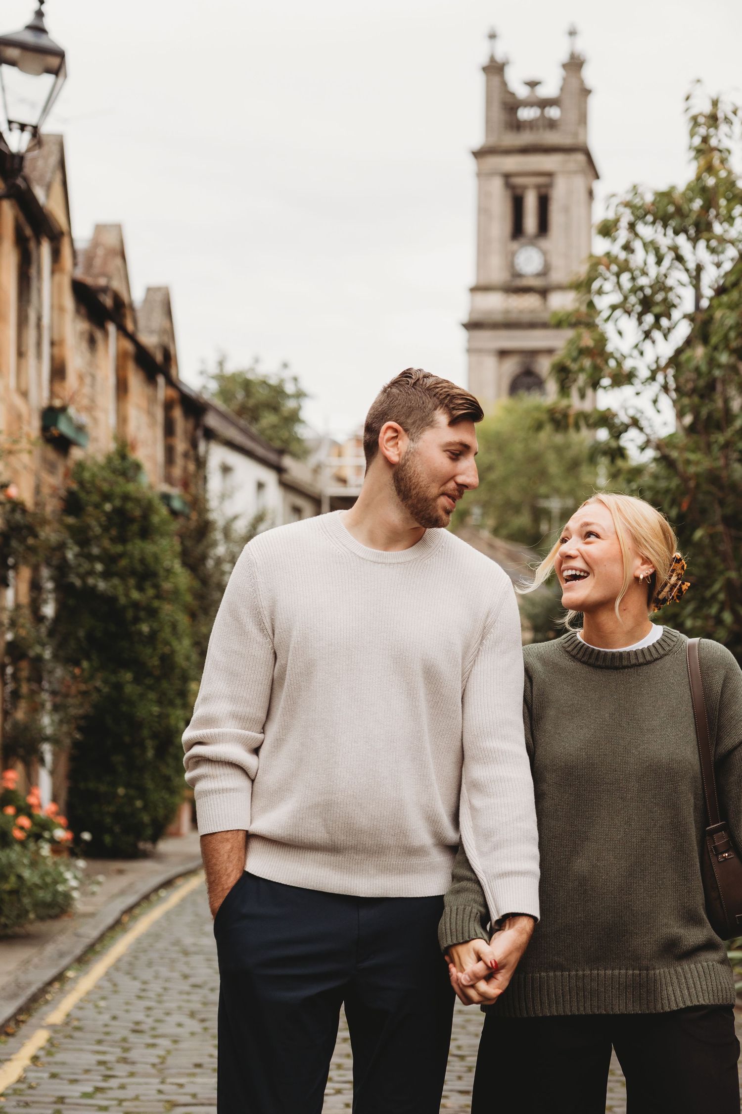A happy couple holds hands and smiles while strolling down a charming cobblestone street with a historic clock tower.