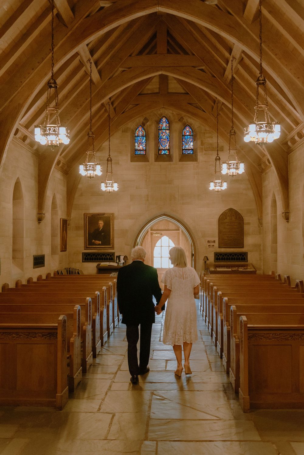 Elderly couple holding hands walking down church aisle toward stained glass windows in a beautiful stone chapel.